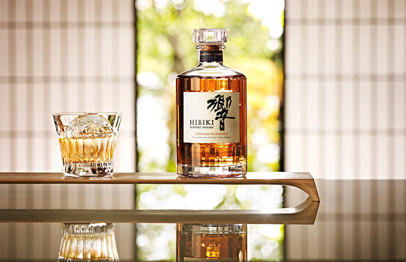 Hibiki Flasche mit einem Cocktail auf einem Holzbrett, das sich in einem Glastisch spiegelt.