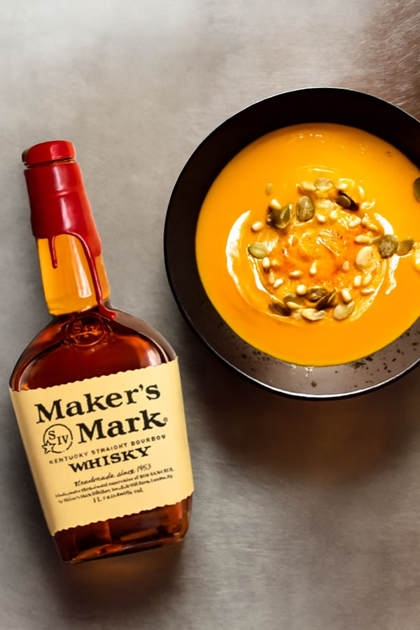 Eine volle Flasche Maker's Marks neben einem Teller gefüllt mit Kürbissuppe, garniert mit Sonnenblumen- und Kürbiskernen