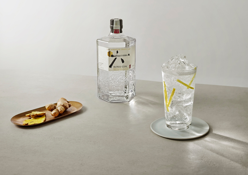 Roku Gin & Tonic im Glas mit Eis und Ingwerstäbchen auf einem grauen Tisch mit eine Falsche Roku Gin und aufgeschnittener Ingwer auf einem Tablett