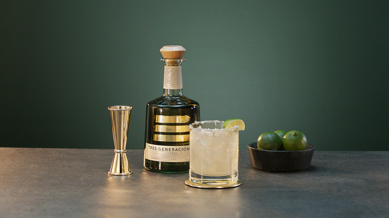 Cocktail Tres Margarita in einem Glas vor einer Flasche Tres Generaciones Plata, einem Messlöffel und einer Schale Limetten
